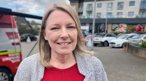 Rob Trigg/BBC Photo of Healy looking into the camera and smiling. She has shoulder-length blonde hair and is wearing a red top with a grey blazer over the top. Out of focus, behind her, is a three-storey building with a row of parked cars in front of it. The sky is cloudy with patches of blue sky. 