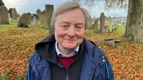 Warwick Lightfoot pictured in front of the graves in the cemetery. He is wearing a red blazer and black hoodie zipped up. He is wearing a blue coat over the top.