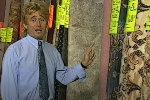 A screengrab from a Mike's Carpets advert. Mr Smith can be seen pointing towards a roll of carpet.