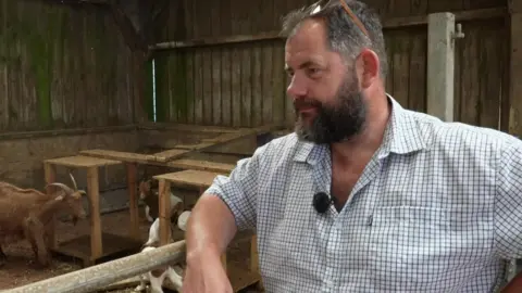 Shaun Ferkin is wearing a white shirt with a pale blue check. He has a dark beard and has sunglasses on his head and is leaning over an enclosure with goats in it. A brown goat with horns on its head and two white goats are in the shot.