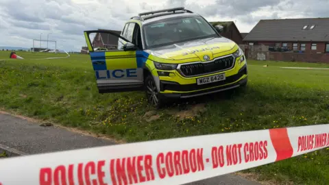 Pacemaker A close up of the red and white police tape cordon which says "Police Inner Cordon, Do Not Cross". Behind it, abandoned on a grassy bank in a housing estate, is the stolen police vehicle. The driver's door of the yellow, blue and white liveried vehicle is lying open