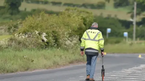 Pacemaker A man in a safety vest with the letters FSNI on his back walks down a street carrying a measuring device.