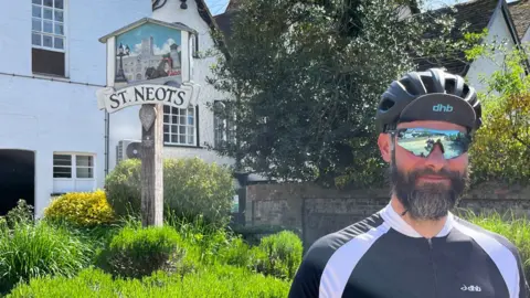 Richard Slade is standing in front of a grassy back with the town sign of St Neots behind him. He is wearing a tight-fitting blue and white cycling top, a bicycle helmet and has sunglasses on. He has a greying beard and moustache.