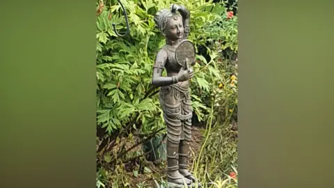 Cambs Police A bronze statue of an Indian woman is placed among shrubs in a garden. She is designed as if wearing ornate clothing and looking at herself in a mirror.