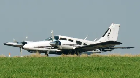 National Crime Agency White twin-engined Piper Seneca aircraft on the ground at an airfield. It is mostly white with some black trip and the identification number N248LT. The grass of the airfield is visible in the foreground. 