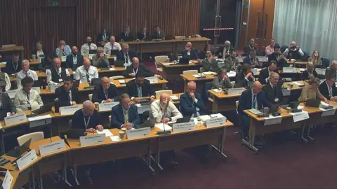 North Northamptonshire Council Lots of people sit behind wooden desks in a large room. 