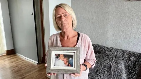 A woman in her fifties with blonde hair and a light pink top is holding a picture.  It shows her as a younger woman hugging her dad.  She is standing in her living room in front of a sofa.