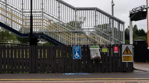 West Midlands Railway A grey footbridge with a metal mesh railing and yellow handrails spans across a train platform. Various signs, including one for "mobility assistance", are visible on a dark wooden fence below the bridge. 
