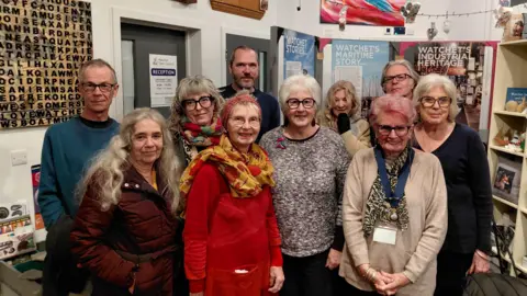 Local Democracy Reporting Service A group of seven women and three men of different ages pose in a community space. Most are smiling. There are posters with the titles "Watchet stories", "Watchet's Maritime story" and "Watchet's industrial heritage" in the background.  