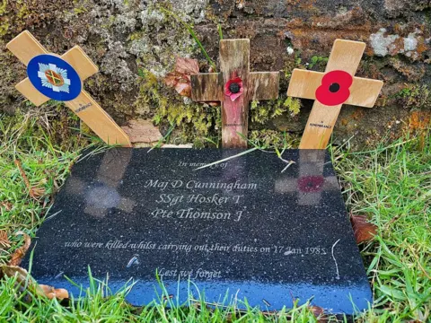 The plaque reads: Maj D Cunningham, S Sgt Hoskar T, Pte Thomson J – who were killed in the line of duty on 17 January 1985, lest we forget. Behind the plague are three small wooden crosses.