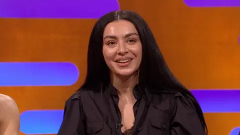 Charli XCX smiles. She has long black hair and wears a black shirt 