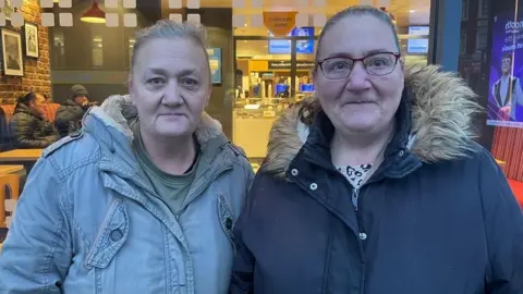Gina Mordan on the right Maria Jones on the left. Ms Mordan wears red framed glasses, a black jacket with brown fur along the collar. Ms Jones wears a khaki jacket with a furry inline hood and a green t-shirt underneath
