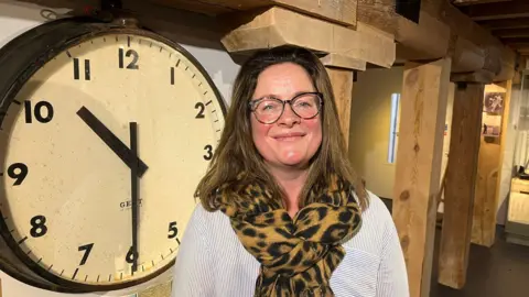 Alison Gudgeon stands in a room next to a large clock showing the time as being 10:30. She has straight, brown hair over her shoulders and wears glasses and a large patterned scarf.