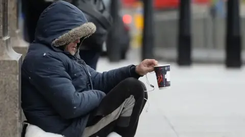 A man crouched against a wall to the left, holding a paper cup and wrapped up in a navy blue coat with a fur lined hood up over his head.