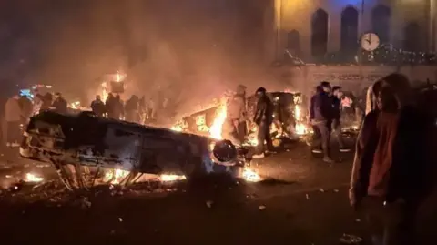 Protesters dressed in black stand around in the dark beside an overturned car on fire.