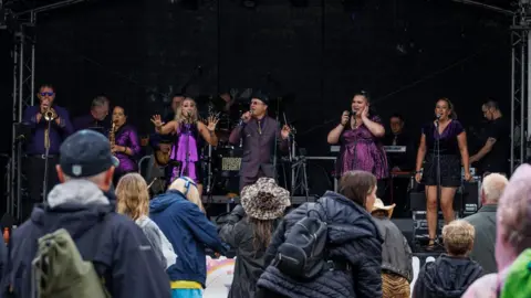 Oak Photography The view from the crowd as a band - Madison Heights - play on a stage with a black background. Four women and two men dressed in purple stand in a line on the front of the stage. From the left, a man is playing a trumpet, a woman is playing saxophone, the others are singing into microphones. Behind them are five more band members but they are not clearly visible - they include a keyboard player and a drummer. The people in the crowd, viewed from behind, are wearing coats and hats.