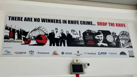 A black, white and red poster is on the wall above a door at Meadowhall. It shows images of Sheffield including Meadowhall, the Winter Garden and Kelham Island Museum