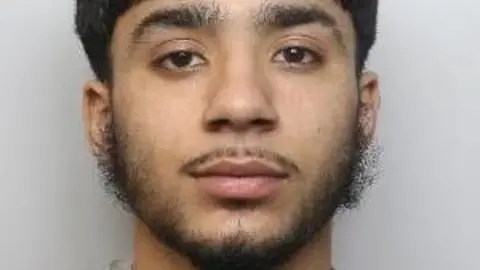 South Yorkshire Police A young Asian man with thick dark hair, a thin moustache and straggly beard looks into the camera.