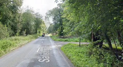 Google A main road through a forested area with a turning on the right and cars both approaching and driving away from the camera