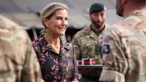 The Duchess of Edinburgh smiling at a soldier in a parade with her hair ted back and hands clasped together