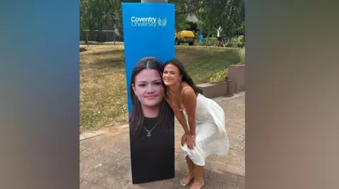 Jennifer Marlow A blue Coventry University banner on a lamp post with Jennifer Marlow's face on and Jennifer Marlow crouched down next to it on the right smiling looking at the camera wearing a white dress with no shoes.