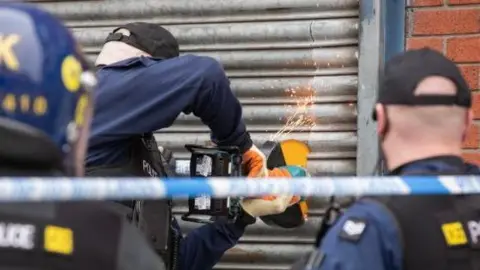 Police are carrying out a raid on the property on Lockett Street. Three police officers are facing towards a metal roller shutter - one is using angle grinder with sparks flying into the air. 