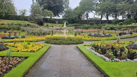 A public garden that has a circular floral display in the middle, with a statue in it. Surrounding it are green spaces with various flowerbeds, they are filled with brightly coloured flowers. In the background is a perimeter hedge and some trees.