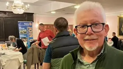 Ian Nightingale, who is wearing a green jacket and shirt and has black glasses, standing in a room where free PSA testing is taking place.
