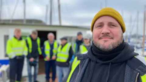 William Gilroy wears a yellow woollen hat and a black top. He is standing on the deck of a boat. six people stand behind him in high visibility jackets. Masts of other boats are visible in the background.