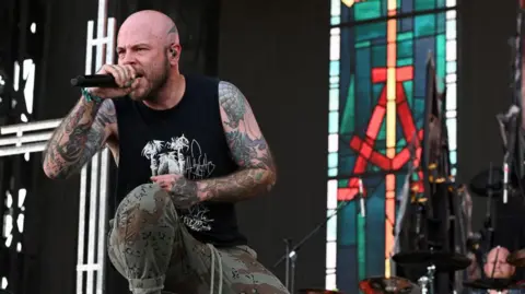 Picture of Ryan Clark, who is the lead singer of metal band Demon Hunter. He is crouched on stage wearing a black vest and camouflage trousers and is holding a microphone.