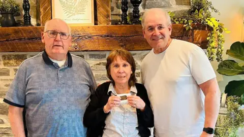 Bridgette Daily and her husband, next to Donald Madar on the right, wearing a white T-shirt. Bridgette in the middle is smiling holding up the dog tags. Her husband has a blue polo shirt on and wears glasses.