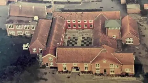 Aerial view of The Red House at Buxton, Norfolk, featuring a quadrangle of red brick buildings and ancillary buildings. There is a chequerboard pattern in the centre of the quadrangle, and greenery to the lower left of the image, and hard standing on all other elevations.