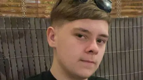 Harvey Willgoose, who has short fair hair and brown eyes. He is wearing black sunglasses on his head.