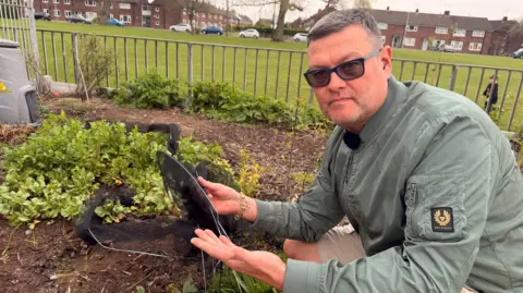 BBC/Jasmine Lowe Tony Henderson - a man in a khaki green coat. He is leaning down with glasses on next to a vandalised vegetable patch. He is holding netting that would have protected the crops from birds, but the netting has been vandalised. 
