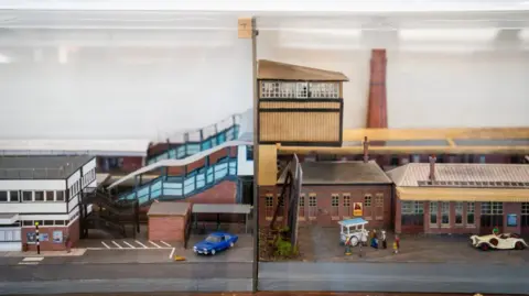 Great Western Railway An image of a model split into two parts: On the left is Gloucester Railway Station in modern times showing the main white building and the steps taking passengers over the line. On the right is the station from bygone years. There is an old-fashioned Rolls Royce-type car at the front.