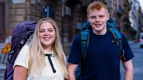 Katie wears a white t-shirt and blue backpack. She has long blonde hair and Harrison has short ginger hair and wears a blue t-shirt and blue backpack with a red hook on the front 