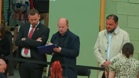 STEVE HUNTLEY/BBC Gavin Callaghan is on the left wearing a dark-coloured suit and red tie. A man with a clipboard is central. And Sam Journet, in a cream jacket, is on the right. Both are looking downwards. The photo is taken from an upper level.