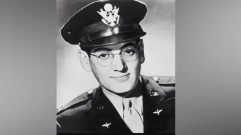 PA Media Glenn Miller pictured in black and white, wearing an army uniform with silver buttons on the shoulders. He is also wearing a hat with a large emblem on the front, and a pale shirt and tie. He had metal framed round glasses and is smiling with his mouth closed and head cocked slightly to one side.