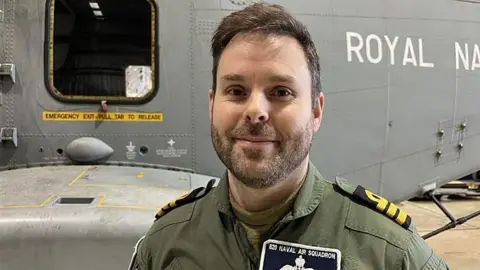 Dressed in a green shirt with a name label Commander Alasdair "Al" Woodward CO of 820 Naval Air Squadron stands in front of a Merlin, like the aircraft which flew to Cyprus.