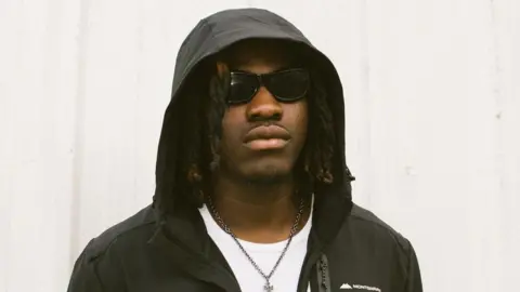 Able Jack is wearing a dark jacket with the hood up and covering his head. He is wearing sunglasses and a white t-shirt with a silver chain