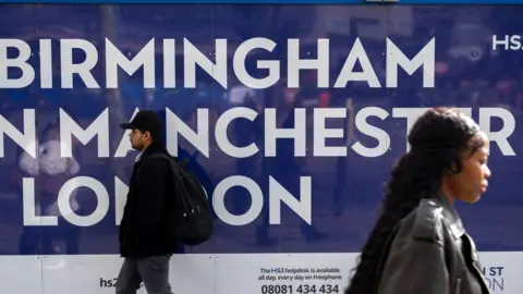 A blue sign with white writing reads "Birmingham Manchester London" as a man and a woman walk past in opposite directions in front of it