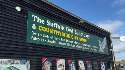 George King/BBC A large black building with a green sign on it which reads The Suffolk Owl Sanctuary and Countryside Gift Shop. There are also pictures of owls, other birds, and a meerkat. 