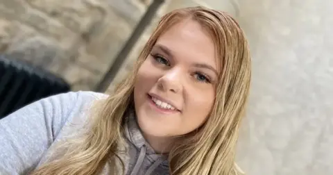 Avon and Somerset Police Selfie of Bethany Branson, a teenager with long blonde hair, blue eyes and long eyelashes. She is wearing a grey hoodie and the background, a wall, is blurred.