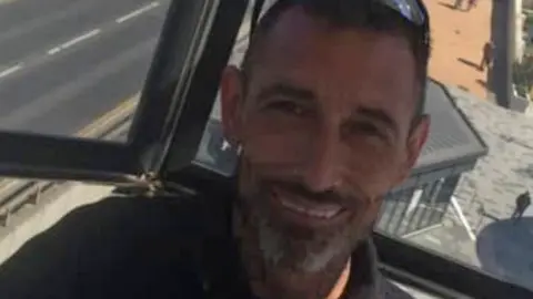 A smiling Adam Lucas has short hair and grey beard and has sunglasses balanced on his head.