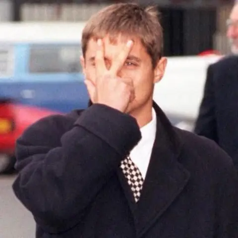 PA Media Un giovane John Alford fa un segno a V davanti al viso con la mano destra. Indossa un elegante cappotto pesante, una camicia bianca e una cravatta a quadri.