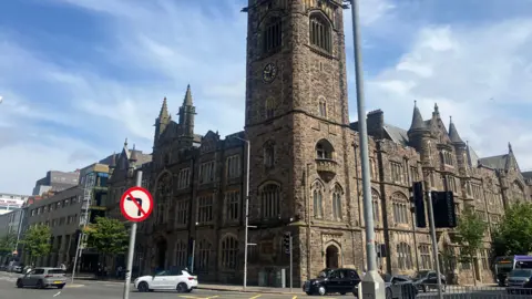 The exterior of the Presbyterian Church House in Belfast. It is on a corner site and is a brown/black/grey bricked imposing building with turrets and towers. there are cars and buses on the road outside.