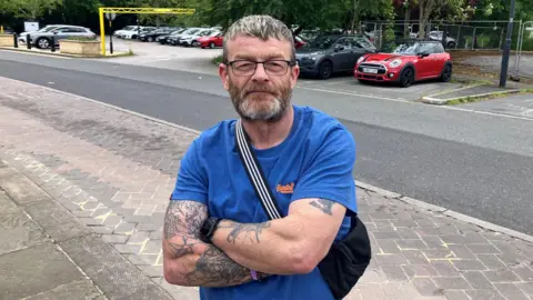 BBC/RICHARD EDWARDS This image shows minicab driver Michael Berry looking straight at the camera. He has his arms folded and has tattoos on his forearms. He is wearing a blue T-shirt, has a beard, short brown hair with fair highlights and is wearing glasses.