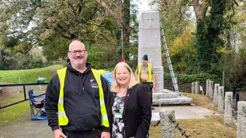 Terri Beer Andrew Stephenson on the left wearing a black hooded jumper with a hi-vis over the top and Terri Beer stood next to him wearing a black blazer and a poppy badge on the right. They are both smiling at the camera. In the background is Steve Davies who is working on the war memorial which has a ladder leaning against it.