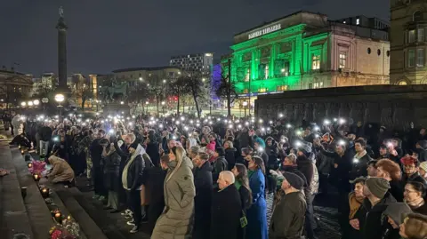 Proudly Said A vigil held for The Vivienne in 2025 with the Liverpool Empire Theatre lit up in green in the background. A group of people pay their respects as they light candles outside St George's Hall in Liverpool.
