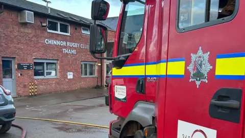 Oxfordshire Fire and Rescue Service A fire engine next to one of the entrances of the rugby club, a two-storey red brick building. 